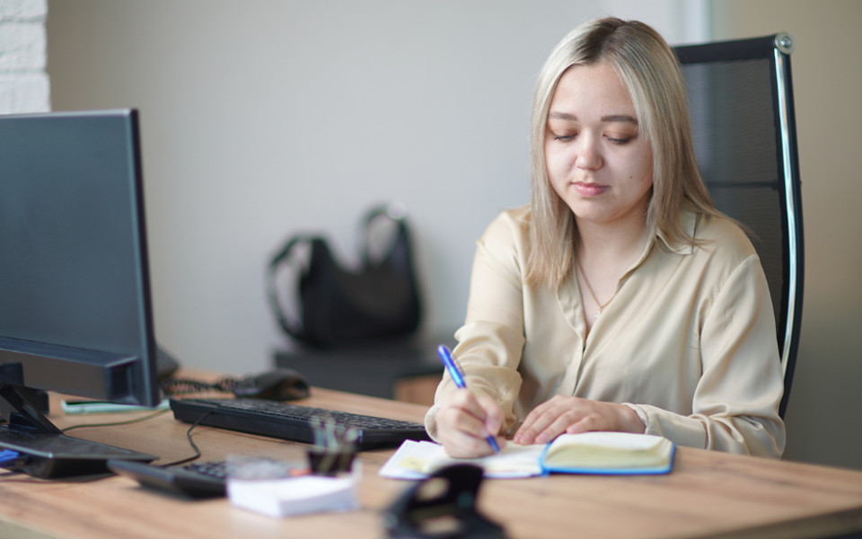 Девушка сидит за компьютерным столом и от руки пишет конспект по базе знаний